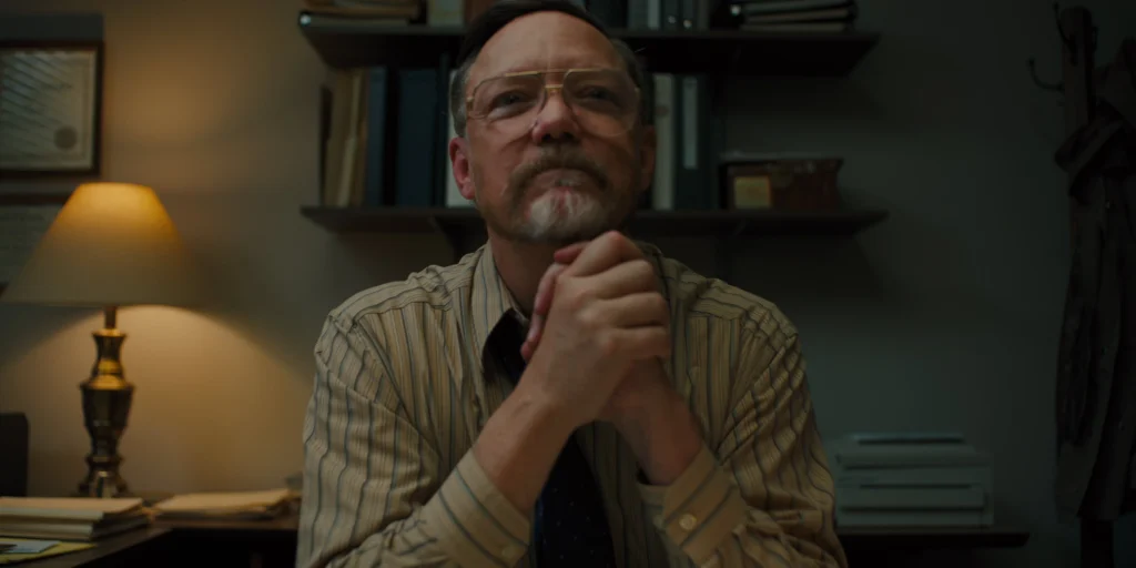 Middle-aged man with glasses and beard, wearing a striped shirt, sits pensively at a desk in a dimly lit room with books and a lamp.