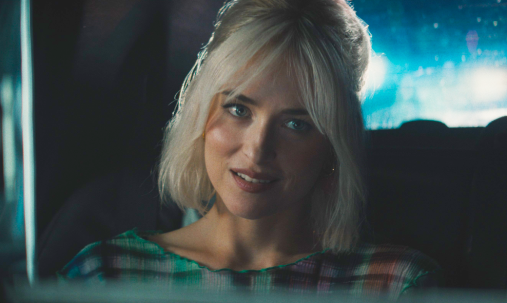 A woman with blonde hair and a checkered top sits in a car, looking slightly to the left.