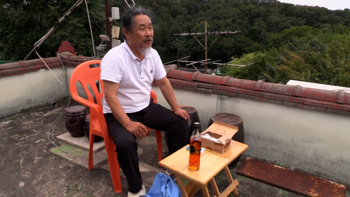 An elderly man with a beard sits on an orange chair on a rooftop. In front of him is a small table with a bottle of liquor, a glass, and a paper bag. Behind him are green trees and antennas.