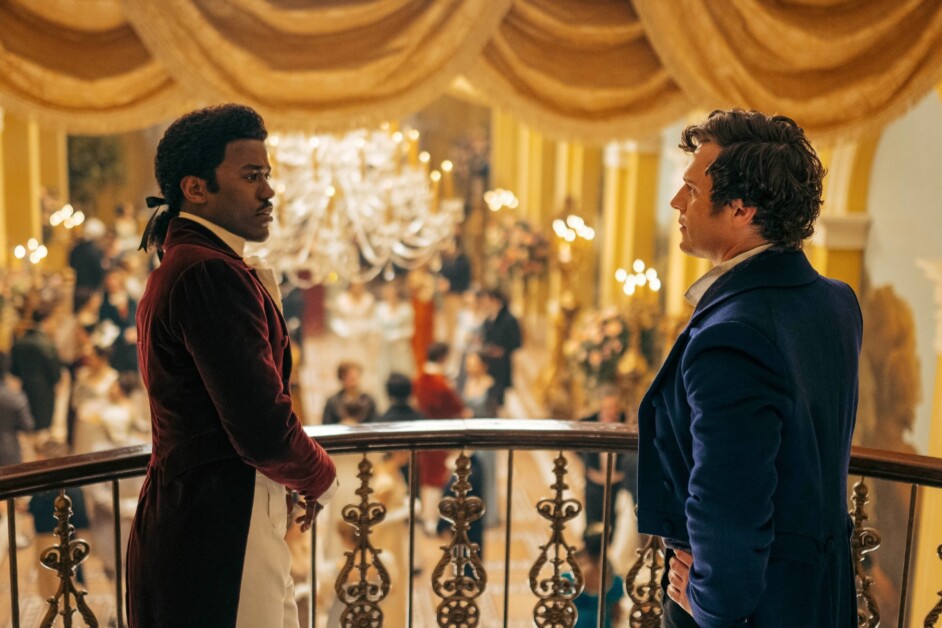 Two men in period costumes stand on a balcony, facing each other during a lively ballroom event. Chandeliers and draped curtains enhance the elegant setting.