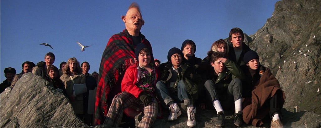 A group of people, including a man with a unique facial appearance, sitting and standing on rocks by the sea, with birds flying in the background.