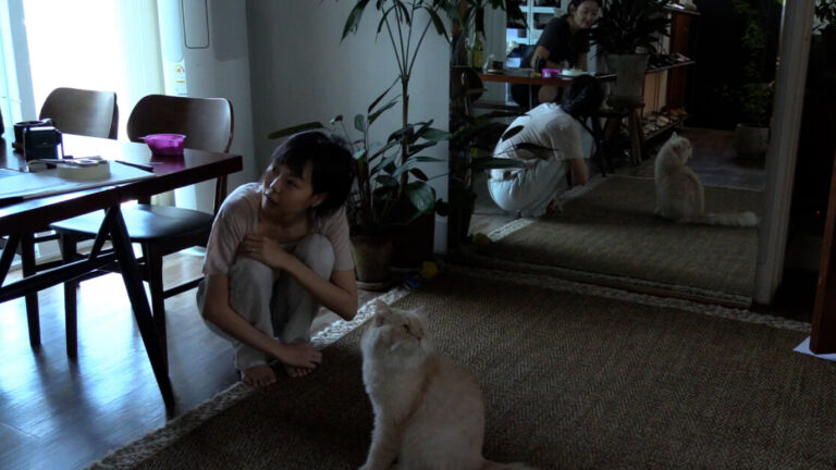 ‘In Our Day’ Review – Quiet Reflection And The Importance Of Cats Inform This Film About Identity Two people and a cat are in a room with wooden flooring and plants. One person is sitting at a table, the other is crouched near the cat, which is looking upwards. A large mirror reflects part of the scene.