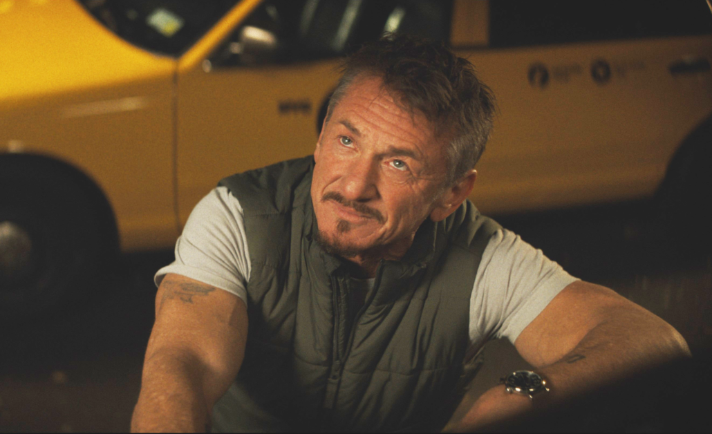 A man with short hair and a goatee, wearing a sleeveless vest over a t-shirt, poses with a slight smile. Behind him, a yellow taxi is partially visible.
