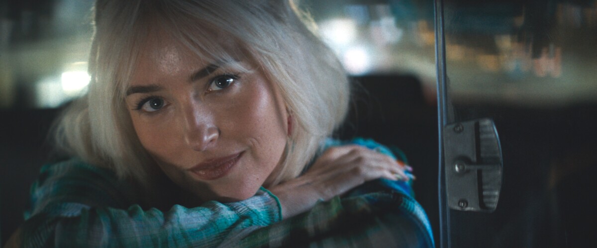 A person with light-colored hair and a plaid shirt rests their arms on the back of a seat and looks forward.