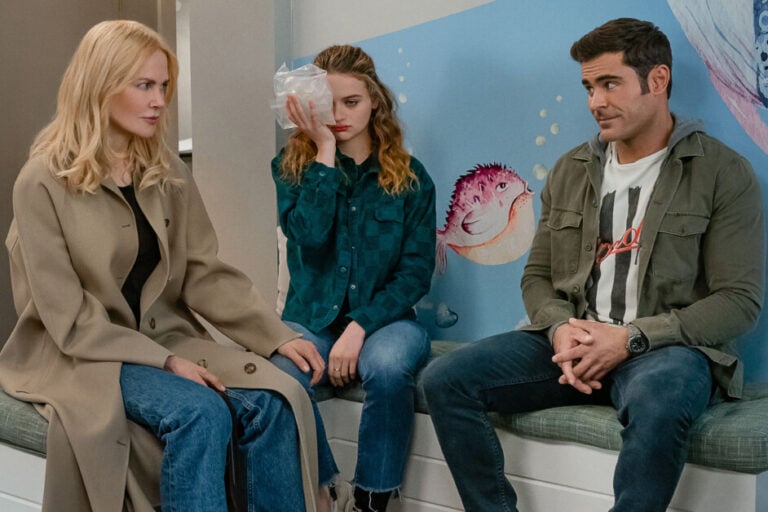 Three people sit on a bench with child-themed wall art behind them. A young girl holds an ice pack to her head, while the woman and man beside her appear to be in conversation with concerned expressions.