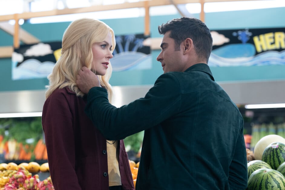 Two people, one with blonde hair and the other with dark hair, are standing close in a grocery store, with the dark-haired person gently touching the blonde-haired person's face. Background shows various produce.