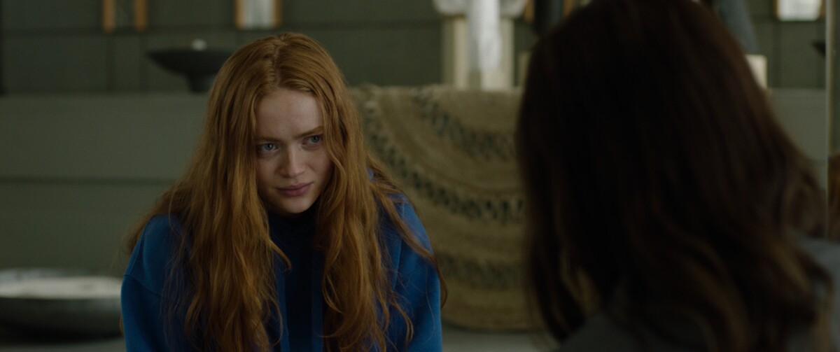 A woman with long red hair, wearing a blue hoodie, looks intently at another person who is partially visible in the foreground. They are indoors with neutral-toned decor in the background.