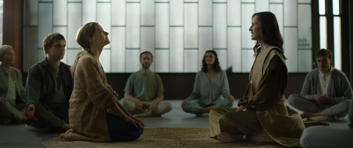 A group of people sit in a circle on the floor, with two women facing each other in the center. They are in a room with large, frosted windows.