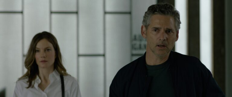 A man and a woman stand indoors with serious expressions, against a background of frosted glass panels.
