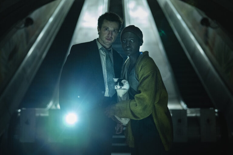 Two individuals stand near an escalator, with one holding a cat. The man on the left wears a suit, while the person on the right wears casual clothes and a hat, with a flashlight illuminating them.