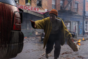 A person in a beanie and yellow jacket, holding a bag and reaching towards a car, runs through a chaotic street with debris and small fires, in front of a damaged grocery store.