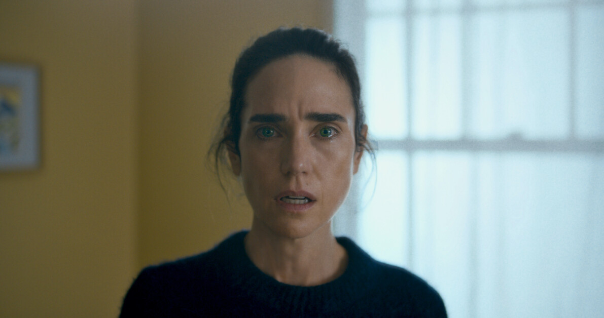 A woman with dark hair looks directly at the camera with a serious expression, standing in a brightly lit room with a window in the background. She is wearing a dark top.