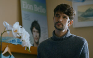 A man with short brown hair and a beard stands indoors wearing a gray sweater. In the background, there is a poster of the same man and a white orchid in the foreground.