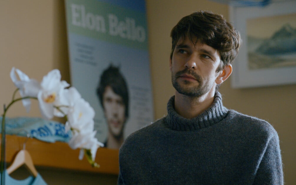 A man with short brown hair and a beard stands indoors wearing a gray sweater. In the background, there is a poster of the same man and a white orchid in the foreground.