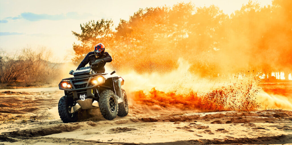 Person riding an ATV through sand, kicking up a large plume of dust, with trees and a sunset in the background.