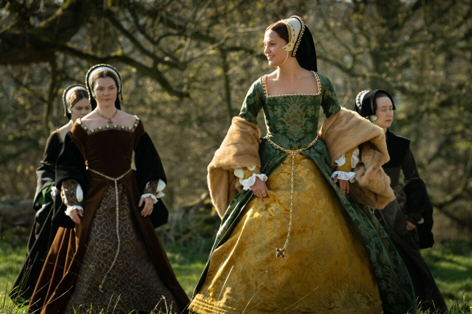 Four women dressed in elaborate, historical costumes walk in a wooded area, with one in a green and gold gown leading the group. Trees and grass are visible in the background.