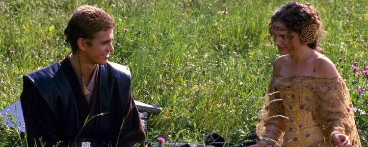 Two people sit on the grass in a meadow, smiling and looking at each other. The person on the left wears dark clothing, and the person on the right wears a patterned dress with elaborate accessories.