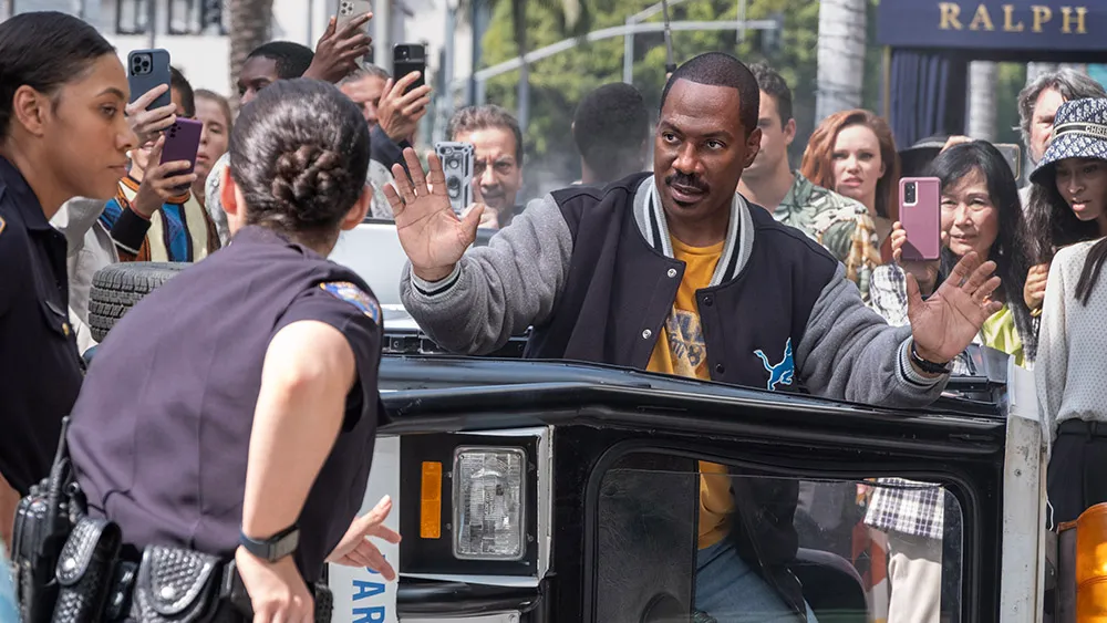 A man raises his hands while exiting a vehicle as police officers and a crowd with phones surround him on a busy street.