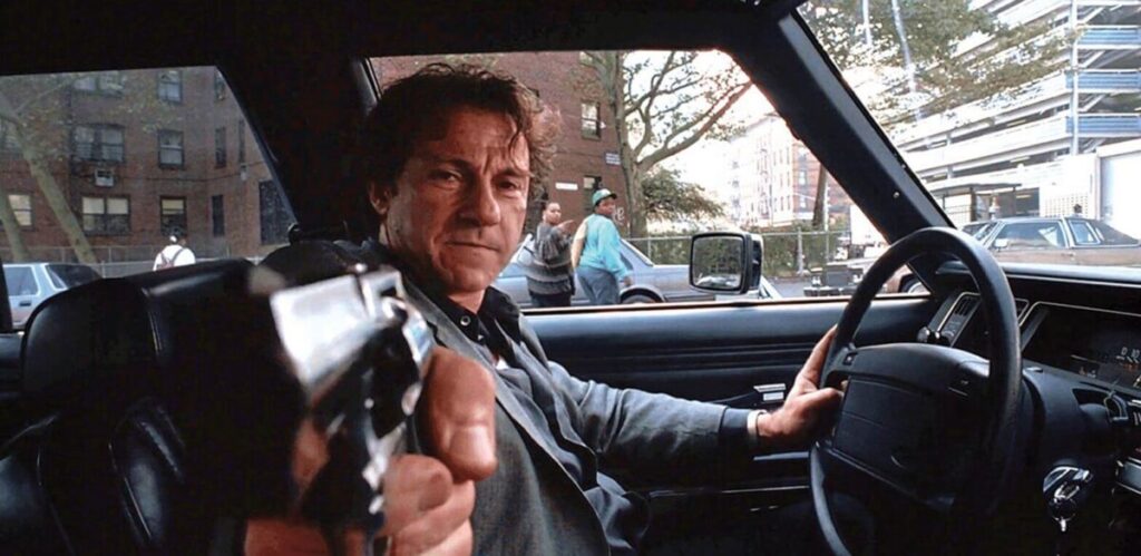 A man in the driver's seat of a car points a gun directly at the camera, with a stern expression. Urban street scene visible through the car window.