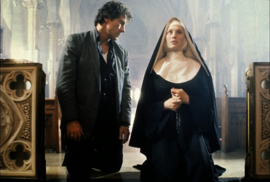 A man and a nun kneel side by side in a church with ornate wooden carvings. The man looks at the nun, who is gazing upwards and holding a rosary.