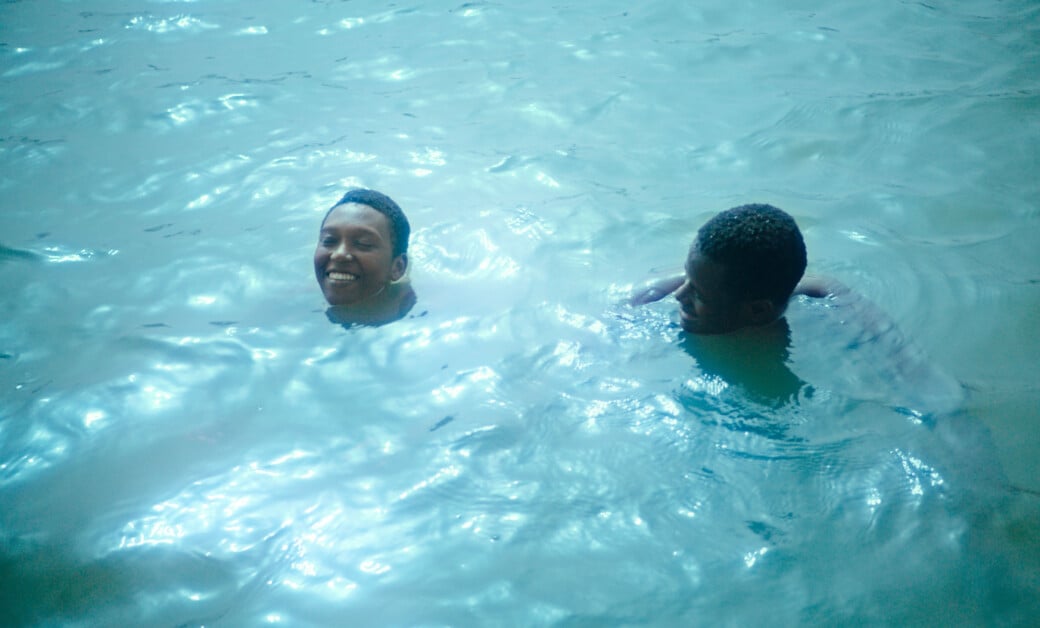 Two individuals are swimming in clear blue water, smiling and enjoying their time. The person on the left is slightly above the water, while the person on the right is partially submerged.