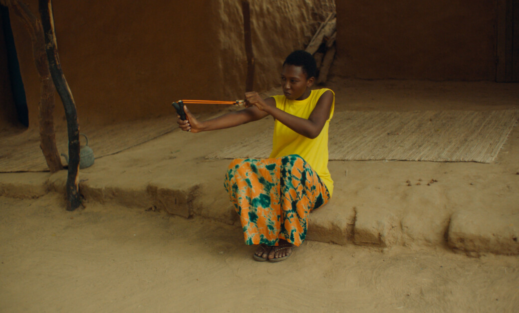 A person in a yellow sleeveless shirt and colorful pants sits on a stone ledge, aiming and pulling back a slingshot.