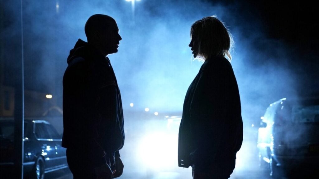 Two people in silhouette stand facing each other in front of bright car headlights, with blue fog and night background.