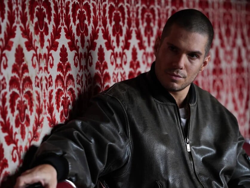 A man with a buzz cut and a serious expression sits against a red and white patterned wall, wearing a leather jacket.