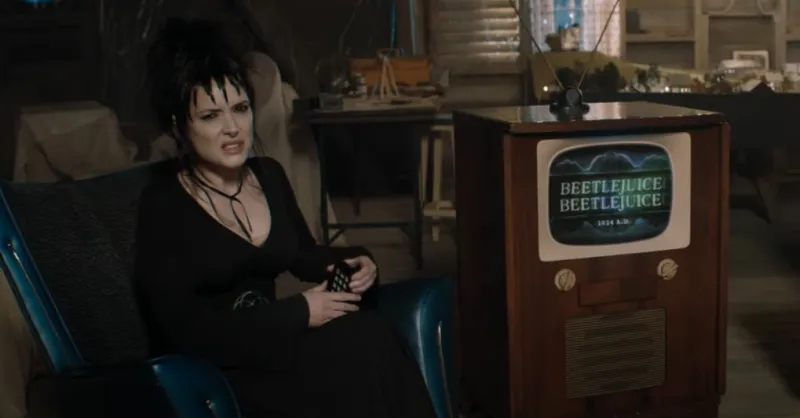 Person in black outfit and dark hair sits in a room with an old television displaying "BEETLEJUICE BEETLEJUICE" on the screen.