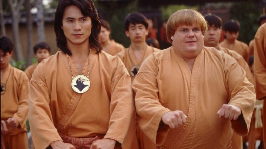 Two men in matching orange martial arts uniforms stand side by side among a group of students in a similar attire, one with long hair and the other with a bowl cut, preparing for practice.