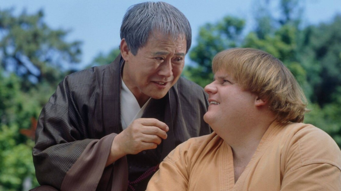 An older man in traditional attire speaks to a younger man with blonde hair who is sitting and listening attentively. They are outdoors with green foliage in the background.