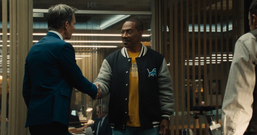 Two men shake hands in an office setting; the man on the left wears a blue suit, and the man on the right wears a black jacket with a light blue logo and a yellow shirt.