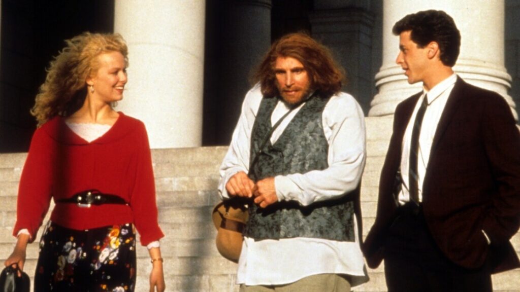 Three people walking down steps. The person in the middle wears a vest and holds a hat; the person on the left wears a red top and floral skirt; the person on the right wears a suit. Stone columns in the background.