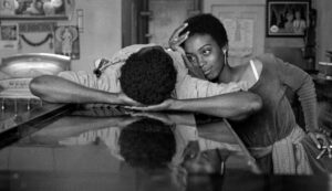 ‘Bushman’ Blu-Ray Review – Forgotten Drama Blurs The Line Between Reality And Fiction A woman gently touches the head of a man resting with his arms folded on a counter in a black-and-white photo. They are in a room with framed pictures and a door in the background.