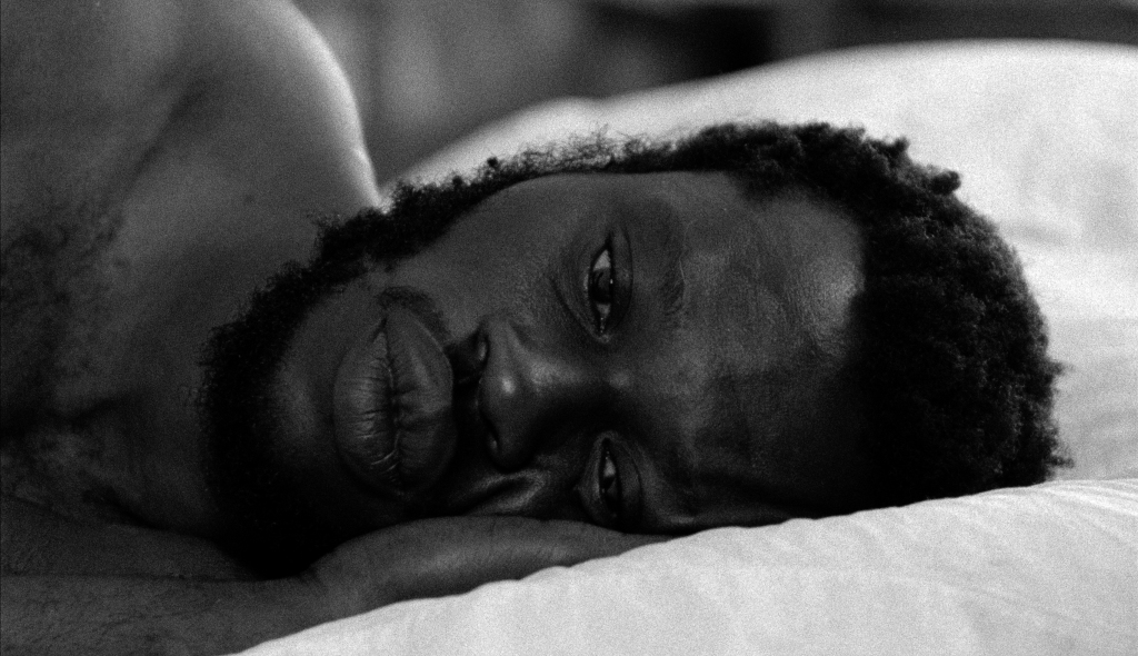 A person with a beard is lying down on a pillow, facing the camera with a thoughtful expression. The image is in black and white.