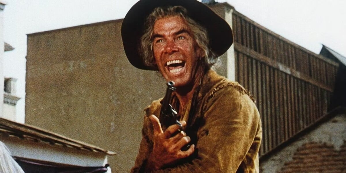 A man wearing a cowboy hat and holding a bottle shouts emphatically outdoors in a Western movie setting.