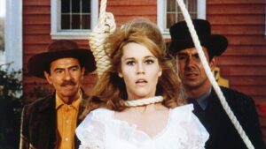 ‘Cat Ballou’ Blu-Ray Review – Western Farce Is An Uproarious Success A woman in a white dress with a noose around her neck stands between two men, one wearing a brown hat and the other a black hat, against the backdrop of a red wooden building.