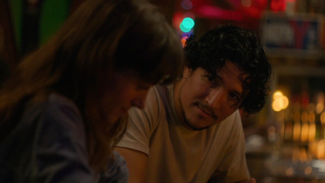 A man with curly hair and a mustache looks at a woman with long hair in a dimly lit setting.
