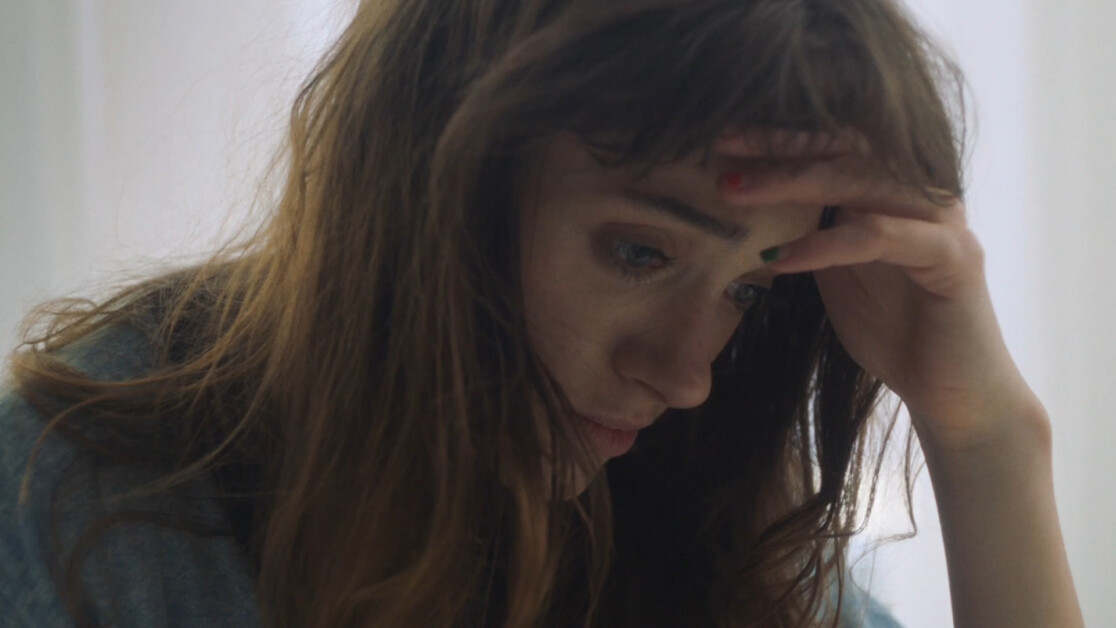 A woman with long brown hair holds her head in her hand, appearing deep in thought or stressed. She is wearing a blue garment and looking downward.