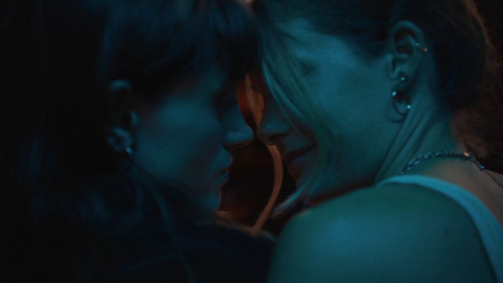 Two people are facing each other closely, illuminated by dim, blue-tinted lighting. One person has long, light-colored hair and multiple ear piercings. Both appear to be engaged in an intimate moment.
