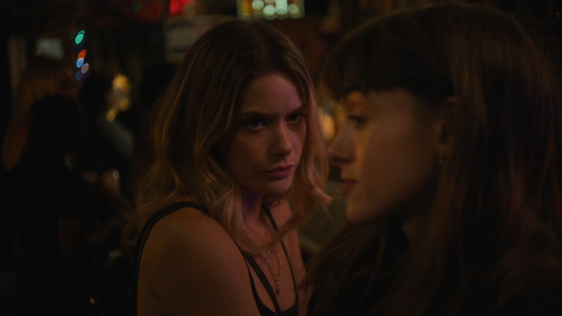 Two women are in a dimly lit indoor setting, one looking directly at the camera with a serious expression while the other looks away. The background is blurred, suggesting a busy environment.