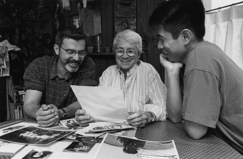 A group of three people, two men and an older woman, sit around a table looking at photographs and smiling. The table is spread with various black and white photos.