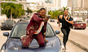 Two men in action: one on a car aiming a handgun, the other running with a pistol. Urban backdrop with palm trees and skyscrapers.