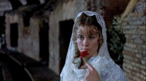 A woman in a white veil holds a red carnation to her lips, standing in front of a stone wall in an outdoor setting.