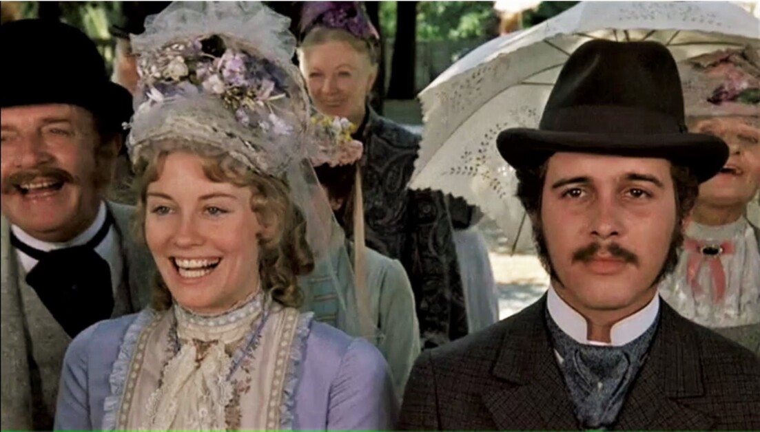 A woman and a man dressed in late 19th-century period costumes smile, standing amid other people. The woman wears a floral hat, and the man wears a dark hat and suit.