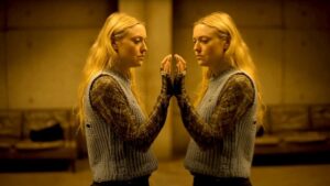 A woman with long blonde hair looks at her reflection in a mirror, touching it with her fingertips. She wears a blue knitted vest over a long-sleeved patterned shirt. The background is dimly lit.