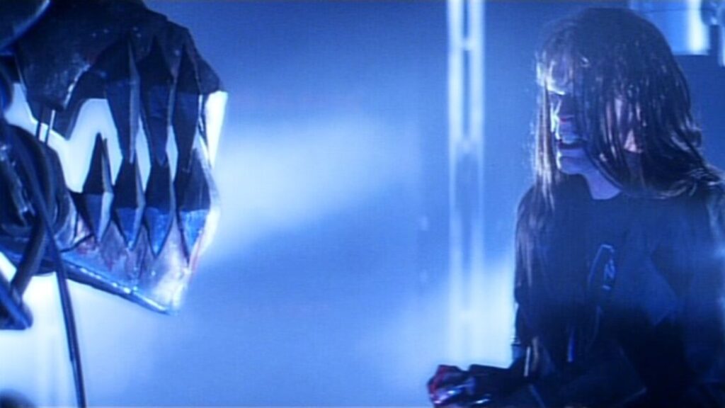 A person with long hair screams while facing a large mechanical creature with sharp teeth in a dimly lit, blue-tinted environment.