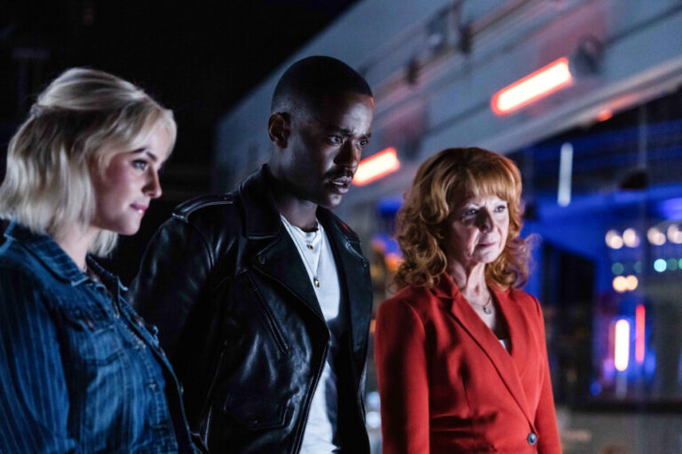 Three people, two women and a man, stand close together indoors, looking intently at something off-camera. The setting is modern with blue and red lighting.
