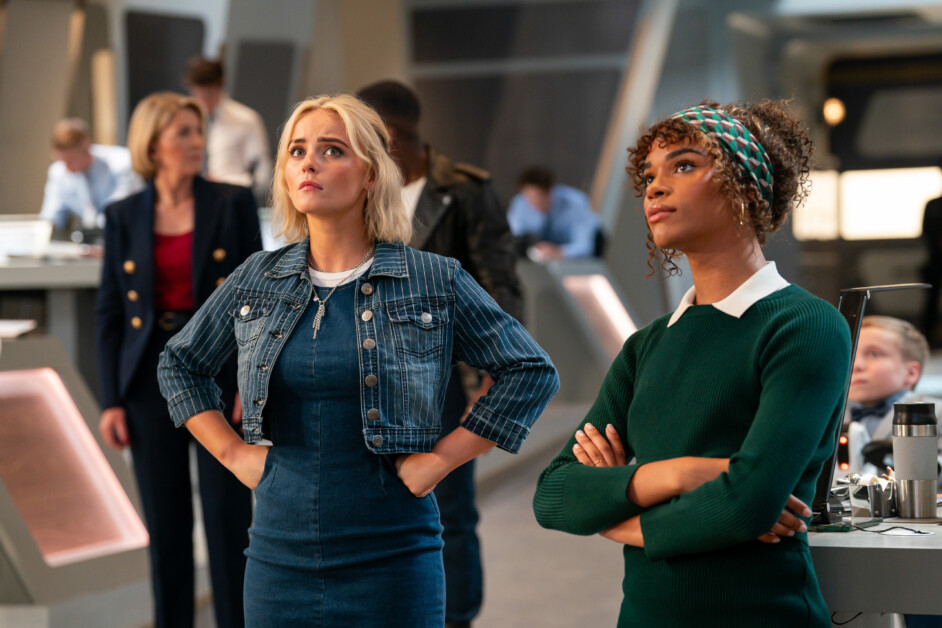 Two women standing side by side, attentively looking upwards, in a busy office setting with people working in the background. One wears a denim jacket and dress, the other a green sweater.