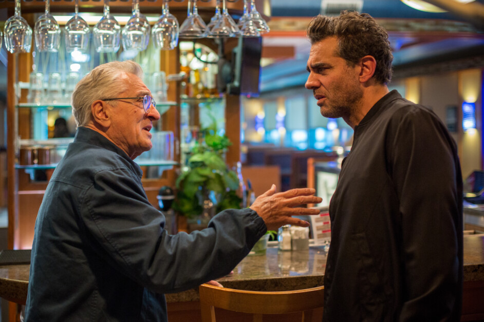 Two men are engaged in a conversation at a bar; one is gesturing while speaking, and the other is listening attentively. The bar area behind them features hanging glasses and various bottles.